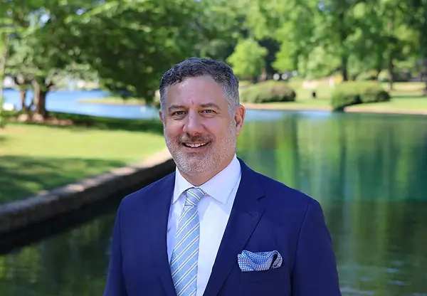 Construction lawyer Gregory L. Sheltong in a blue suit and striped tie standing beside a lake in a sunny park, smiling at the camera.